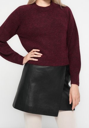 Personne portant un pull bordeaux à manches longues et une jupe en cuir noire, posant avec une main sur la hanche sur un fond uni.