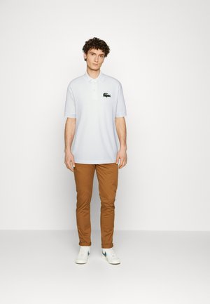 Camisa polo blanca con un logo de cocodrilo negro, combinada con pantalones marrones y zapatillas blancas, con mangas cortas y un cuello clásico.