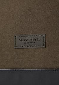 Tissu vert foncé avec une texture lisse, présentant une étiquette en cuir noir sur laquelle est inscrit "Marc O'Polo" et "EST. À STOCKHOLM."