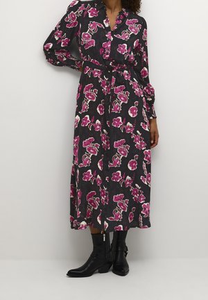 Femme portant une robe midi noire avec un imprimé floral rose vif et blanc, une taille ceinturée, des manches longues, associée à des bottines noires.