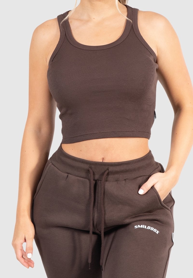 Femme portant un débardeur court marron cintré et un pantalon de jogging marron assorti à cordon, une main dans la poche.