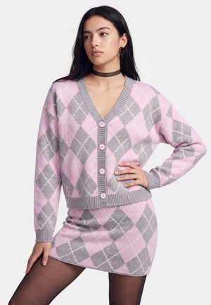 Vrouw die een roze en grijze argyle cardigan en bijpassende rok met zwarte panty's draagt, staand met één hand op de heup tegen een witte achtergrond.