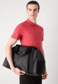 Črn potovalni nahrbtnik z bleščečo površino, z močnimi ročaji, stranskim žepom in vtisnjenim logotipom na vrhu. Drži ga model v rdeči majici.