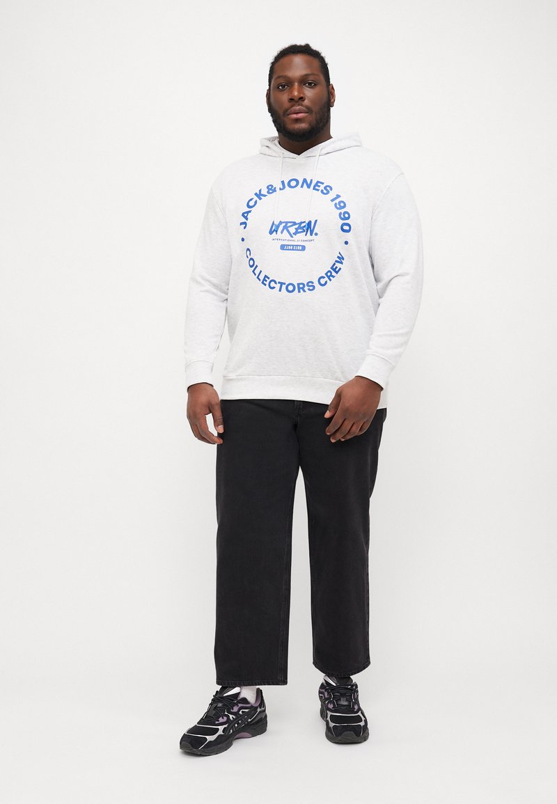 Grauer Hoodie mit blauer Grafikdruck, der ein rundes Logo zeigt. Schwarze, lockere Hose und schwarze Sneakers mit lila Akzenten.