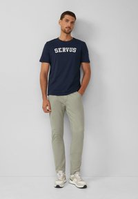 Granatowa bawełniana koszulka z napisem "SERVUS", zestawiona z jasnoszarymi dopasowanymi spodniami oraz beżowymi sneakersami z białymi i szarymi akcentami.