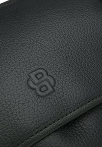 Accesorio de cuero negro con superficie texturizada, logotipo en relieve y detalle de cremallera visible a lo largo del borde.