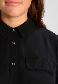 Czarna koszula z guzikami, z kołnierzykiem, gładkiego materiału, z pojedynczą kieszenią na piersi z klapą oraz okrągłymi srebrnymi guzikami.