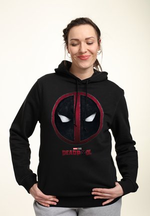 Kvinde iført en sort Deadpool-hættetrøje, hænderne hvilende på hofterne, smilende og kigger nedad mod en lys, ensfarvet baggrund.