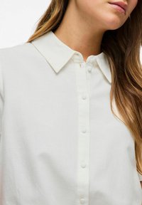 Camicia bianca con colletto classico, tessuto liscio e vestibilità dritta. Presenta una fila di bottoni lungo la parte anteriore.