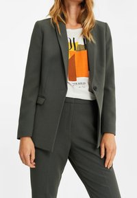 Tmavě zelené na míru šité sako s jedním knoflíkem, se zářezovými klopami a dvěma předními kapsami, doplněné o tričko s potiskem pod n�ím.