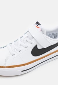 Valkoinen lenkkari mustalla Nike-merkinnällä, vaaleanruskea kuminen välipohja, vaaleanharmaat nauhat ja tarranauha. Rakennekoristeet lähellä pohjaa.