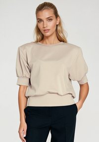 Beżowa bluzka z bufkami, wykonana z miękkiego materiału, z okrągłym dekoltem i ściągaczem na dolnej krawędzi, zestawiona z ciemnymi spodniami. Gładka faktura z minimalnymi detalami.