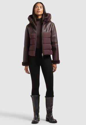 Femme portant une doudoune violette à capuche avec bordure en fourrure, pantalon noir et bottes en cuir noires montant jusqu'aux genoux, debout devant un fond gris.