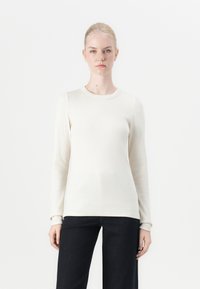 Lange mouwen crème ribbel top, ronde halslijn, zachte textuur, aansluitende silhouet, gecombineerd met donkere wijde jeans.