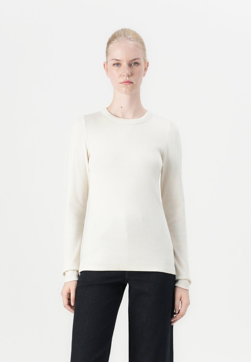 Lange mouwen crème ribbel top, ronde halslijn, zachte textuur, aansluitende silhouet, gecombineerd met donkere wijde jeans.