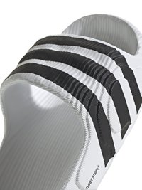 Bela drsna sandal z tremi teksturiranimi črnimi črtami čez zgornji del. Ima gladko, oblikovano podplato in ravno, odprto zasnovo.