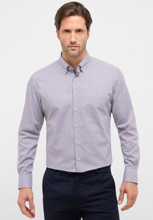 Man draagt een geruit overhemd met lange mouwen en drukknopen, en donkere broek, staand tegen een eenvoudige lichte achtergrond.