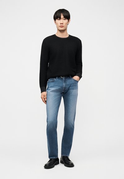 Νέος άνδρας όρθιος με το χέρι στην τσέπη, φορώντας μαύρο πουλόβερ με υφή, ξεθωριασμένο μπλε τζιν και μαύρα παπούτσια, σε απλό φόντο.