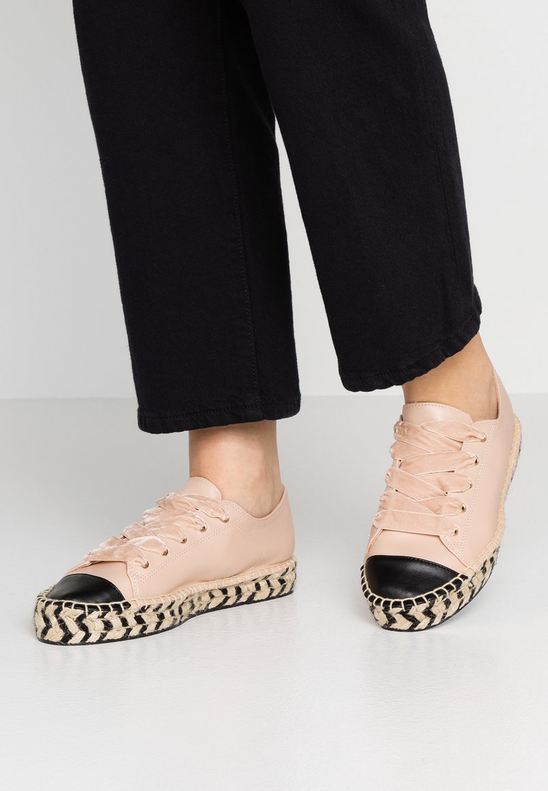 Pieds portant des baskets espadrilles à lacets rose clair avec des bouts noirs et des semelles à motif tissé, associées à un pantalon noir court.