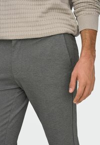 Hallid kitsenevad püksid, mis on valmistatud tekstuurse kangaga, küljetaskutega ja kitsas lõikega. Kätel asetatud reiele, peal helebeež kampsun.