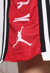 Shorts de sport rouges avec des rayures latérales noires. Présente un logo Jumpman blanc et du texte blanc, fabriqués en matériau léger, finition texturée.