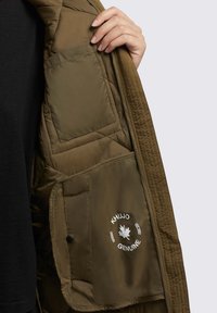 Veste gonflée vert olive à l'intérieur en texture matelassée, poche latérale avec fermeture à pression et détail du logo.