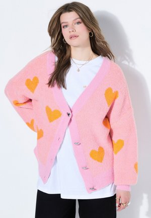 Junge Frau mit langen braunen Haaren trägt einen übergroßen pinken Cardigan mit orangenen Herzen über einem weißen Hemd und schwarzen Hosen und posiert vor einer weißen Wand.