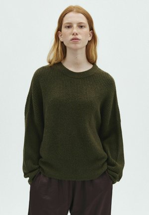 Jeune femme aux cheveux roux portant un pull côtelé vert foncé et un pantalon foncé, debout devant un fond clair uni.