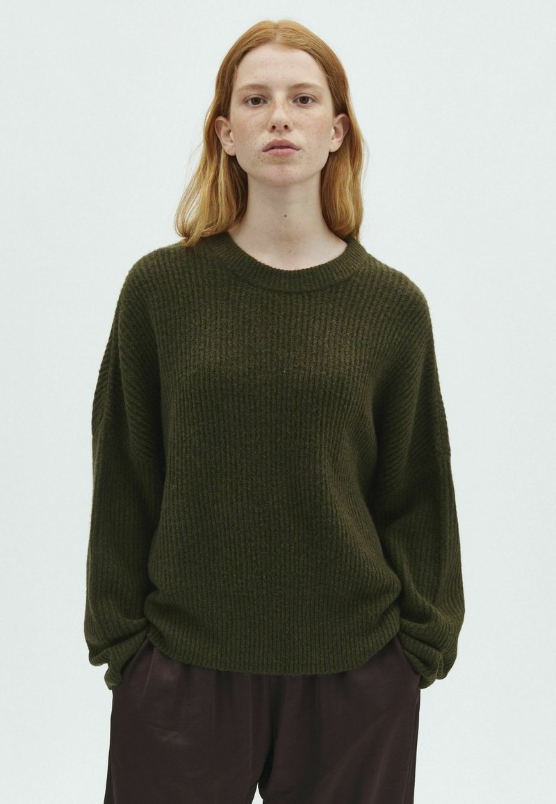Jeune femme aux cheveux roux portant un pull côtelé vert foncé et un pantalon foncé, debout devant un fond clair uni.