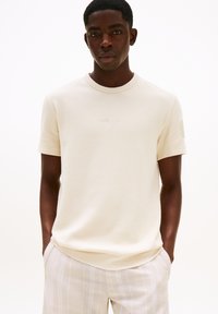 Camisa de hombre de manga corta con canalé en color crema y cuello redondo, con un diseño sutíl en el pecho, combinada con pantalones claros a rayas.