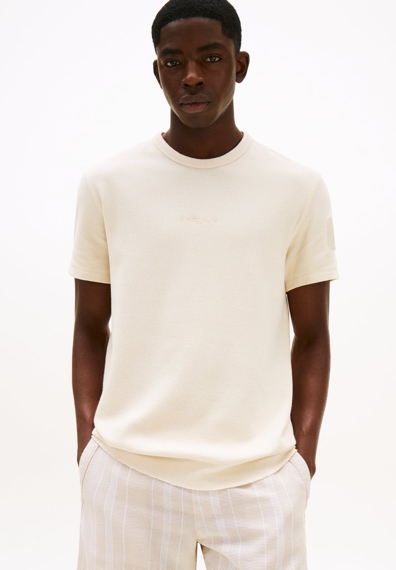 Camisa de hombre de manga corta con canalé en color crema y cuello redondo, con un diseño sutíl en el pecho, combinada con pantalones claros a rayas.