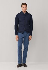 Marineblaue Steppweste mit horizontalen Steppnähten und fünf Knöpfen, über einem kabelgestrickten Langarmshirt, kombiniert mit blauer Hose und braunen Schuhen.