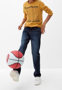 Žlutá dlouhá košile s černým textem, modré džíny a bílé tenisky; osoba kopající červený a bílý basketbal.