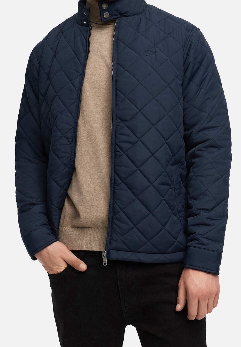 Homme portant une veste matelassée bleu marine sur un pull beige, main dans la poche, et un pantalon noir sur un fond uni.