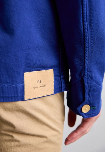 Blue denim jacket with a tan leather patch displaying “PS Paul Smith” and a button closure details on the sleeve.