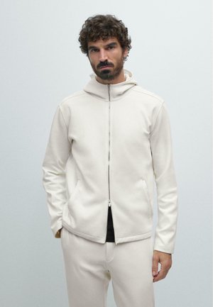 Homme aux cheveux bouclés et barbe, portant un sweat à capuche blanc zippé et un pantalon blanc assorti, se tenant devant un fond clair uni.