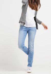 Jean en denim bleu clair à coupe slim, avec des coutures contrastantes et une taille mi-haute. Assorti à un cardigan gris et un haut blanc.