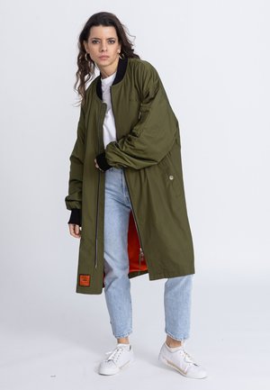 Olive green bomber jacket with a zip closure, featuring black accents, orange lining, and a textured exterior. Worn over a white shirt and jeans.
