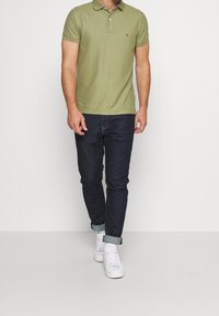 Homme portant un polo vert olive, un jean bleu foncé retroussé, des baskets blanches et des chaussettes blanches, se tenant devant un fond blanc uni.