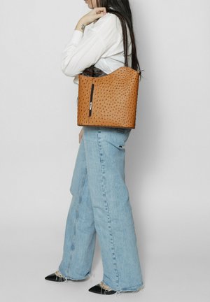 Sac fourre-tout en cuir texturé brun, avec un haut arrondi, une seule bandoulière et une poche zippée à l'avant. Porté avec un jean large bleu.