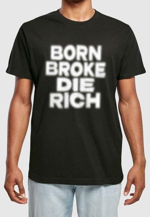 Mies, jolla on musta t-paita, jossa on sumeat valkoiset tekstit "BORN BROKE DIE RICH", yhdistettynä vaaleansinisiin farkkuihin, seisoo yksinkertaisen taustan edessä.