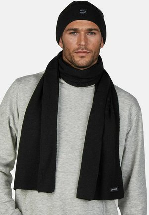 Man draagt zwarte gebreide muts en bijpassende sjaal over lichtgrijze trui, kijkt recht vooruit tegen een eenvoudige lichte achtergrond.