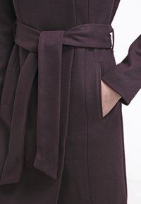 Dlouhý, tmavě fialový kabát se zavazovacím páskem, předními kapsami a hladkým povrchem látky. Má strukturované ramena a rovný siluetu.