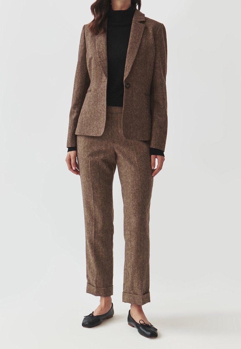 Femme portant un blazer marron ajusté et un pantalon assorti avec revers, un col roulé noir et des ballerines noires, debout devant un fond uni.