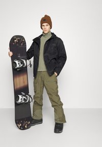 Snowboard con attacchi neri tenuto da una persona in giacca nera, pantaloni verde oliva e un berretto marrone su uno sfondo neutro.