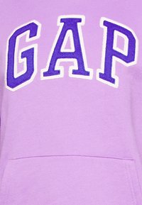 Felpa viola con la scritta "GAP" in grandi lettere ricamate bianche e viola scuro. Presenta una tasca frontale e un tessuto morbido e texturizzato.