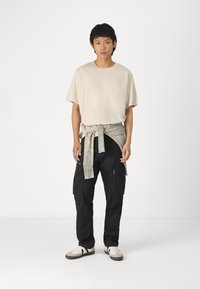 Beige oversized t-shirt, knyttet lys grå stof ved taljen, sorte cargo bukser med lommer, og hvide sko med sorte striber.