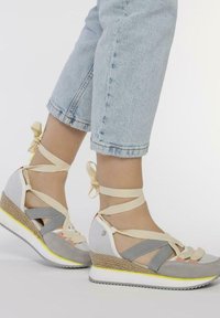Sandales à talon compensé en maille grise et blanche et en daim, avec des accents jaunes et des lacets beiges enroulés autour de la cheville. Talon tressé texturé.