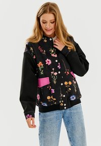 Czarna kwiecista kurtka z wielokolorowymi wzorami, różowe kieszenie, zapięcie na guziki i ściągacze przy mankietach, w połączeniu z jasnoniebieskimi dżinsami.
