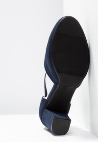 Escarpin en nubuck bleu marine avec un bout pointu, un talon haut et une semelle en caoutchouc noir texturé. Il présente un dos ouvert avec un détail de sangle.